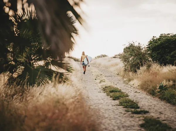 Surfer walking dusty path toward beach